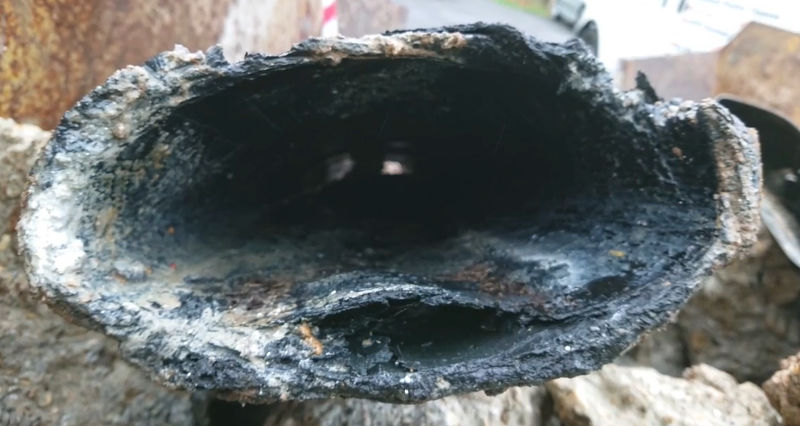 Cut end of a pitch fibre pipe, oval in section, with the inner wall broken up and shedding material into the bore